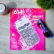 614 magazine August cover, hot pink with can of hard seltzer. Magazine is surrounded by a can of seltzer, a plant, and sunglasses.