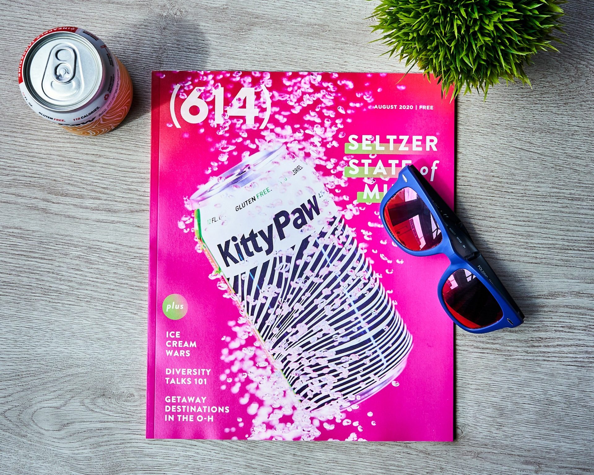 614 magazine August cover, hot pink with can of hard seltzer. Magazine is surrounded by a can of seltzer, a plant, and sunglasses.